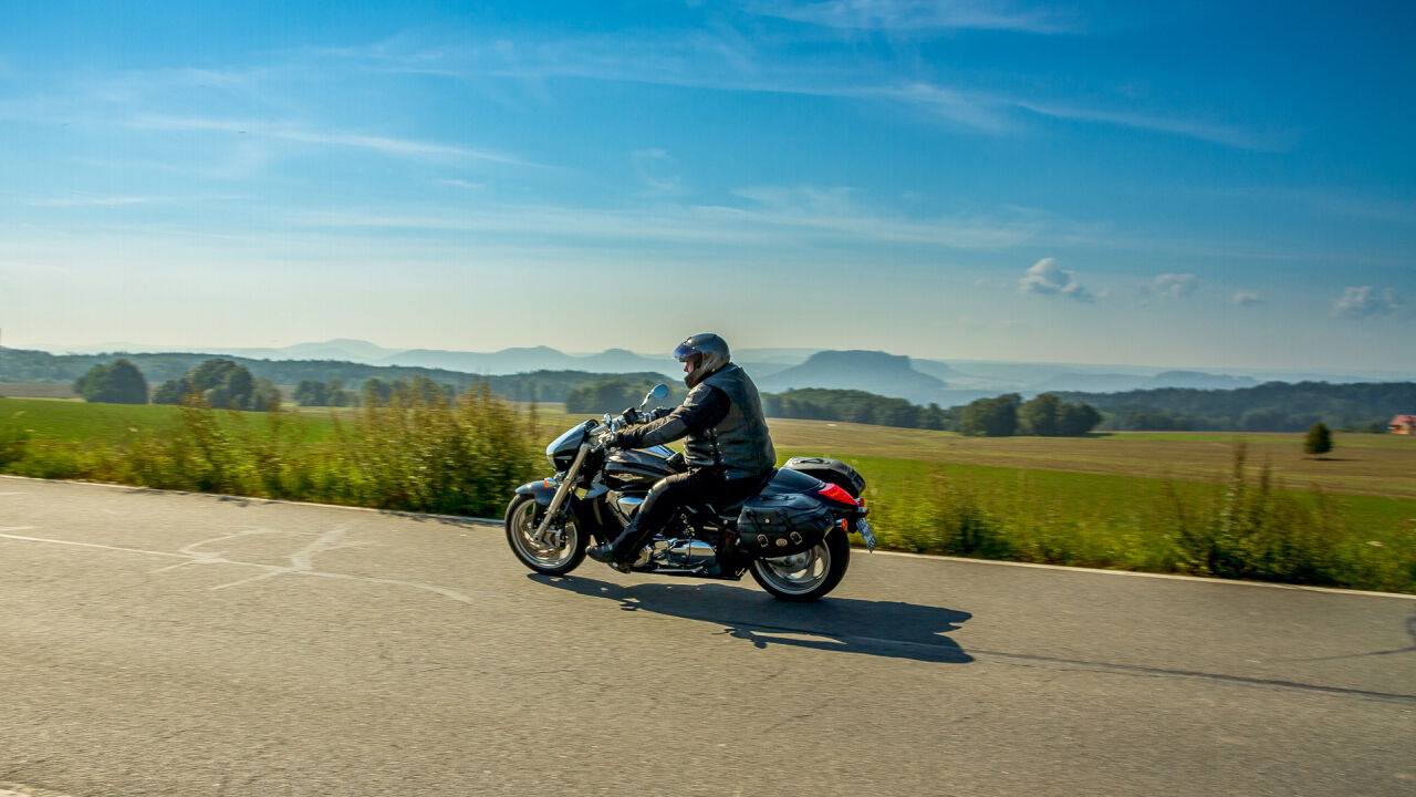Motorradfahrer auf einer Landstraße, dahinter flache Felder und am Horizont Tafelberge