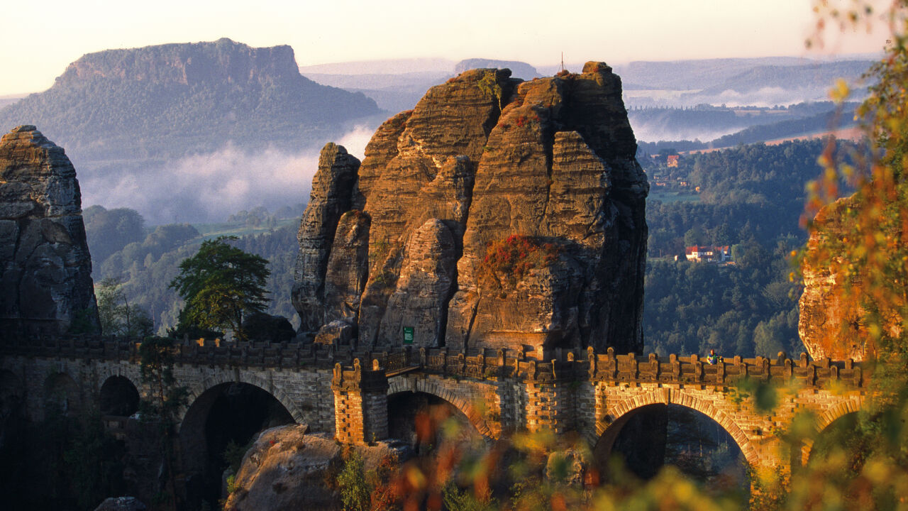 Steinerne Bogenbrücke zwischen hohen Sandsteinfelsen, dahinter ein Tafelberg im Morgenlicht